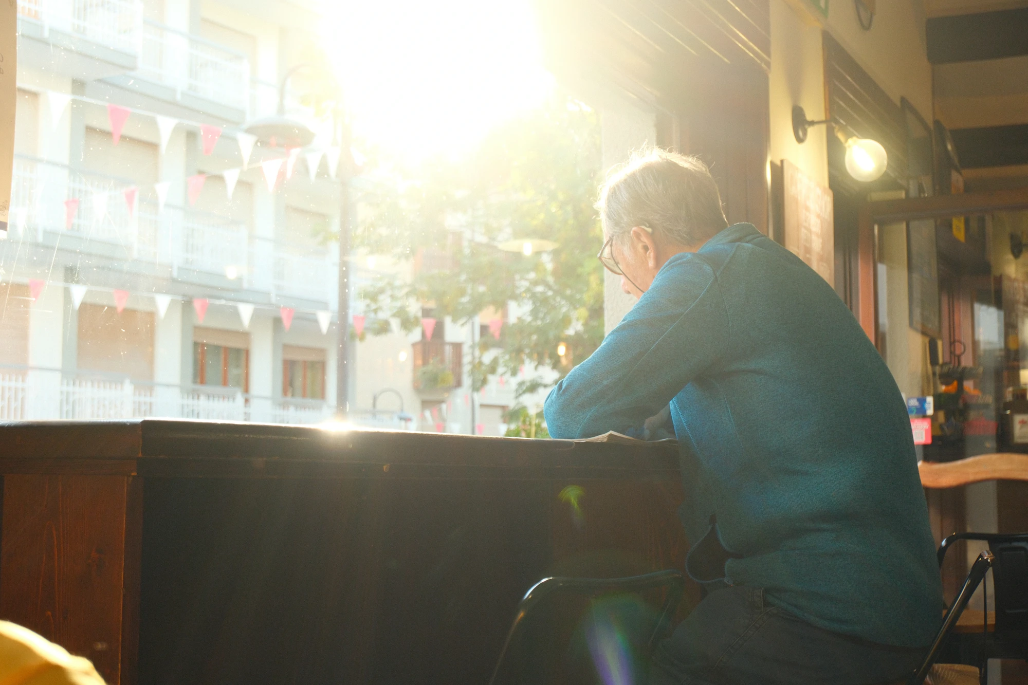 A man sitting at a sunlit café window, reading, with his own street outside